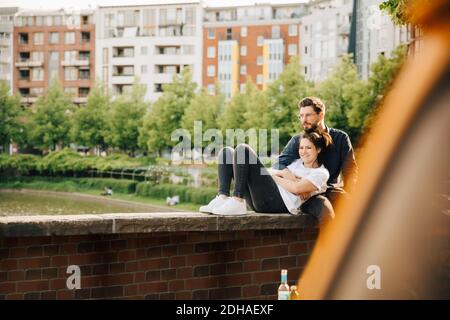 Young man with smiling female partner looking away while sitting on retaining wall Stock Photo