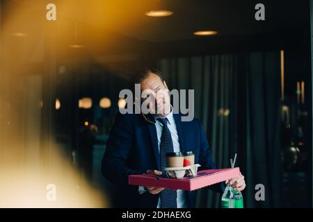 Mature businessman talking on mobile phone while carrying food and drinks in cafe Stock Photo