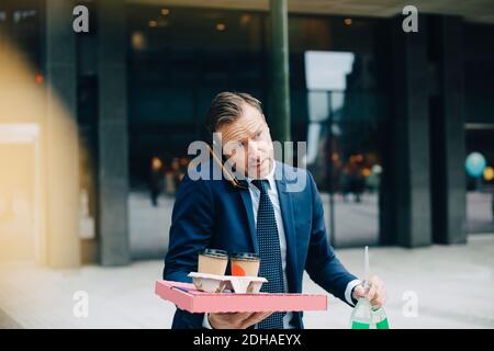 Mature businessman talking on mobile phone while carrying food and drinks in city Stock Photo