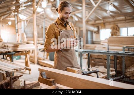 Portrait of a handsome carpenter standing with a smart phone, having a brake during the woodwork in the workshop Stock Photo