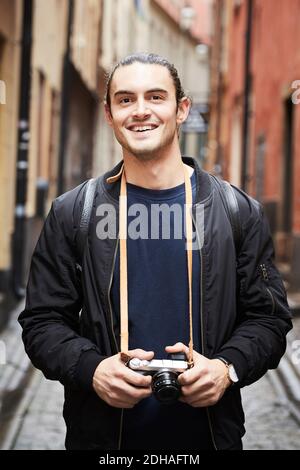 Portrait of smiling young man holding camera while standing in alley Stock Photo