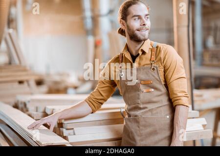 Portrait of a cheerful carpenter standing near the raw wood materials at the carpentry shop warehouse Stock Photo