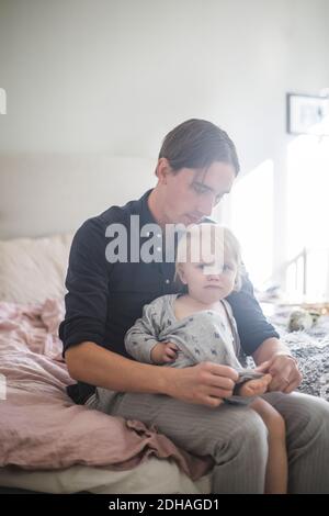 Father assisting baby boy wearing clothes while sitting on bed at home Stock Photo