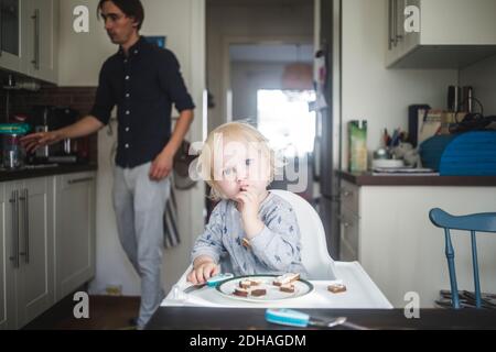Portrait of blond baby boy eating while sitting on high chair at kitchen with father in background Stock Photo