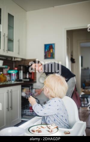 Blond baby boy sitting on high chair while looking at father in kitchen Stock Photo