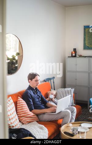 Man using laptop while sitting by toddler son drinking milk from bottle on sofa in apartment Stock Photo