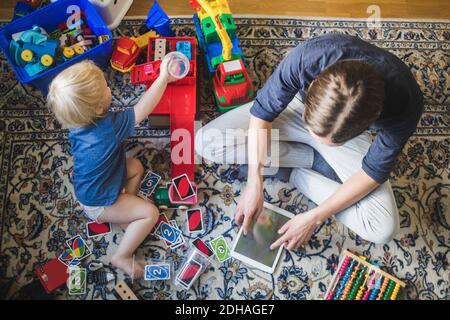 High angle view of blond baby boy playing toys by father using digital tablet in living room at home Stock Photo