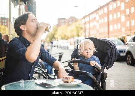Baby boy looking away while sitting on stroller by father drinking at sidewalk cafe in city Stock Photo