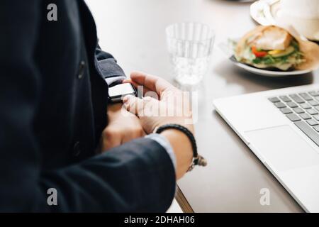 Midsection of businessman checking smart watch while sitting with laptop at desk in office Stock Photo
