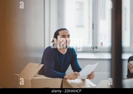 Young businessman holding document while leaning on cardboard boxes in new office Stock Photo
