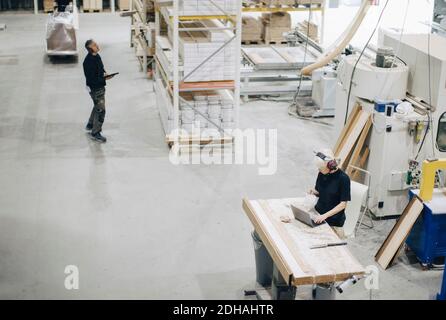 High angle view of male worker examining planks on rack while colleague using laptop at industry Stock Photo