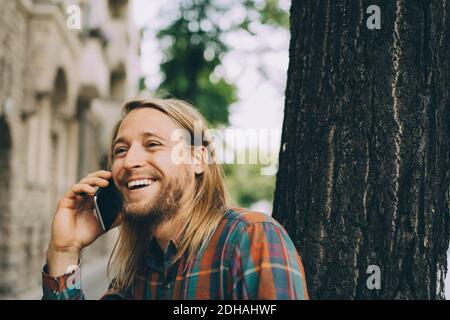 Smiling man talking on mobile phone while leaning on tree trunk in city Stock Photo