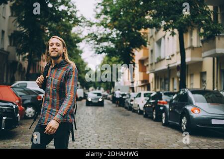 Young man carrying backpack while walking on cobbled street amidst cars in city Stock Photo