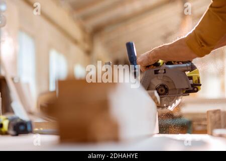 Carpenter sawing wooden bars with cordless electric saw at the joiner's workshop. Close-up with no face Stock Photo