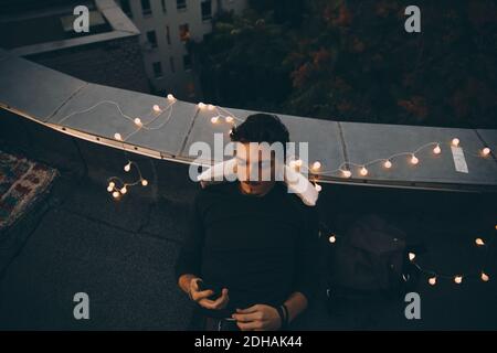 High angle view of young man using smart phone while relaxing on illuminated terrace at dusk Stock Photo