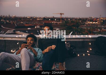 Thoughtful man sitting with friend using mobile phone on terrace in city Stock Photo