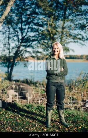 Full length portrait of female farmer standing arms crossed on organic farm with pig in background Stock Photo