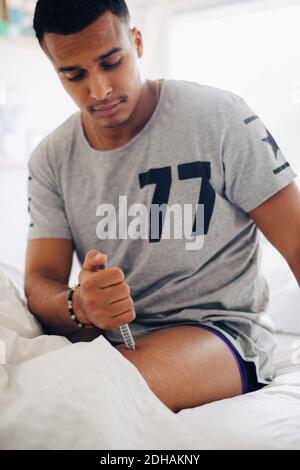 man sitting on bed while injecting insulin on lap at home Stock Photo