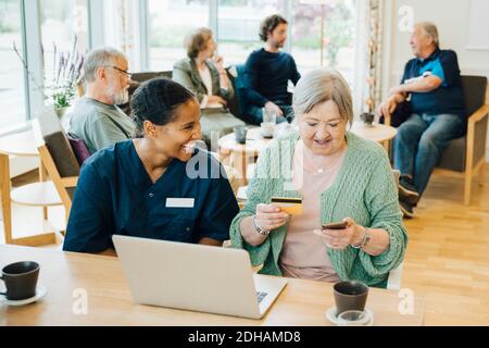 Cheerful female healthcare worker sitting by senior woman doing online shopping with credit card and smart phone in nurs Stock Photo