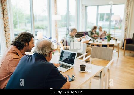 Man assisting elderly grandfather using laptop at dining table in retirement home Stock Photo
