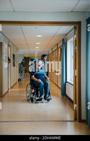 Full length of female nurse pushing disabled elderly man on wheelchair while looking away in alley at retirement home Stock Photo