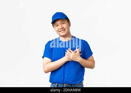 Touched cute asian courier in blue uniform love his clients, care for customer parcels, holding hands on heart and smilig flatte Stock Photo