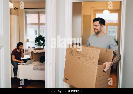 Smiling man carrying box from doorway towards woman using laptop in bedroom Stock Photo