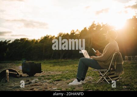 Full length of thoughtful man with laptop and mobile phone sitting on chair during sunset Stock Photo