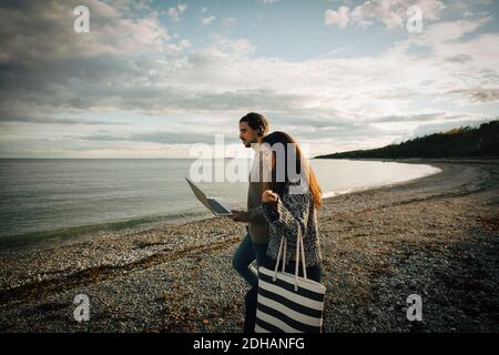 Woman walking with man using laptop on sea shore at beach against sky Stock Photo