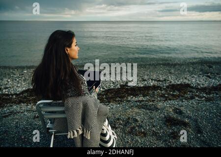 Thoughtful woman with laptop sitting on chair at beach during sunset Stock Photo