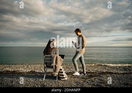 Man using mobile phone while woman sitting with laptop on chair at beach against sky Stock Photo