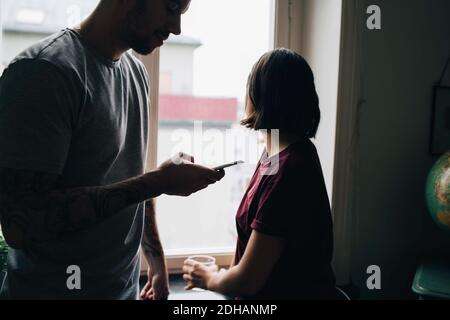 Woman looking through window while man using mobile phone at home Stock Photo