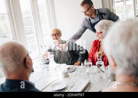 Owner serving water to smiling senior man sitting with friends in restaurant Stock Photo