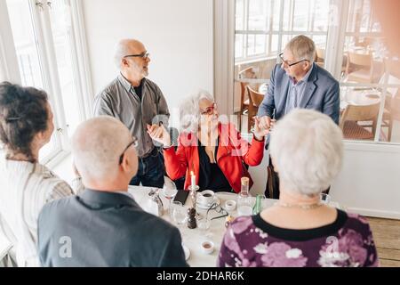 Senior men holding hands with senior woman while friends standing by table in restaurant Stock Photo