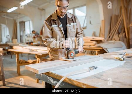 Handsome carpenter in uniform brushing wood with hand machine at the carpentry manufacturing Stock Photo