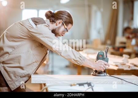 Handsome carpenter in uniform chamfers wooden bar with a hand machine at the carpentry manufacturing Stock Photo