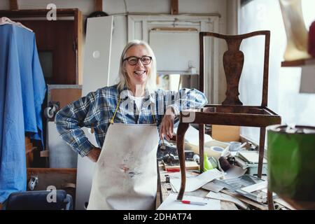 Portrait of smiling female standing by wooden chair at workbench Stock Photo