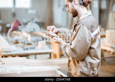 Handsome carpenter working on wooden carvings using a smart phone at the carpentry manufacturing. Concept of using mobile technologies in carpentry Stock Photo