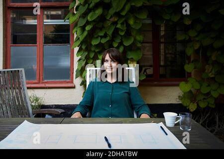 Portrait of confident female architect with blueprint at table in backyard Stock Photo