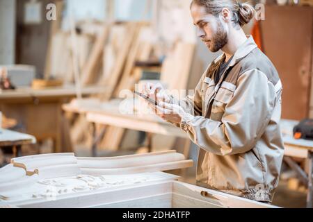 Handsome carpenter working on wooden carvings using a smart phone at the carpentry manufacturing. Concept of using mobile technologies in carpentry Stock Photo
