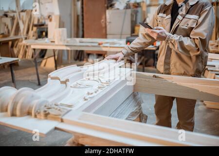 Handsome carpenter working on wooden carvings using a smart phone at the carpentry manufacturing. Concept of using mobile technologies in carpentry Stock Photo