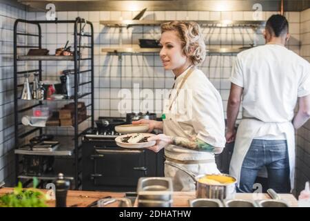 Side view of confident waitress carrying food plates in kitchen at restaurant Stock Photo