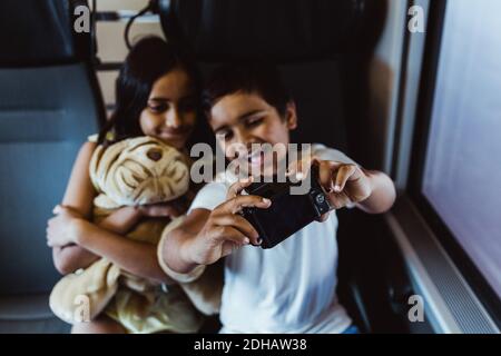 Happy boy taking selfie with sister on camera while traveling in train Stock Photo