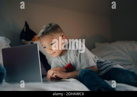 Boy using laptop reclining with mobile phone on bed at home Stock Photo
