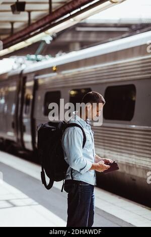 Mid adult man with backpack using mobile phone while standing by train on platform Stock Photo