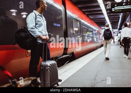 Male tourist with luggage looking away while standing against train on railroad station platform Stock Photo