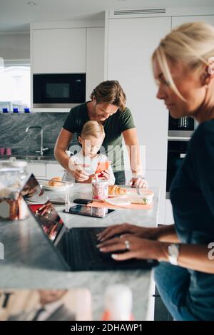 Woman using laptop while man and son holding smart phone in kitchen at home Stock Photo