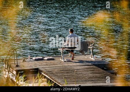 Rear view of man using laptop while sitting on bench over jetty against lake Stock Photo