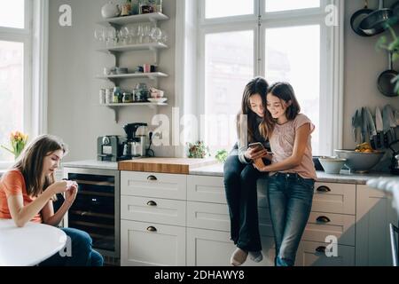 Female friends using mobile phone in kitchen at home Stock Photo