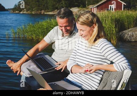Smiling couple using laptop while sitting on bench against lake Stock Photo